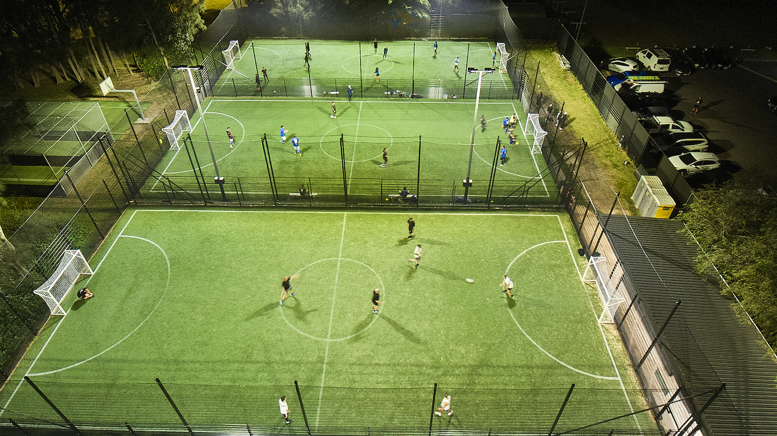 Two young players chasing a ball inside a caged 5-a-side pitch at a KIKOFF venue on a weeknight.